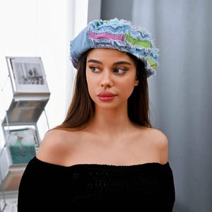 Könnte beinhalten: Eine Jeans-Baskenmütze mit Patchwork-Design, ausgefransten Kanten und Akzenten aus rosa und grünem Stoff. Die Mütze besteht aus hellblauen Denim-Einsätzen und zeigt ein strukturiertes, abgenutztes Aussehen mit sichtbaren Nähten und offenen Kanten.
