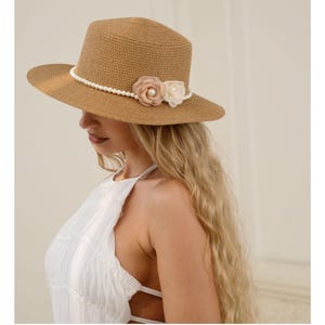 May include: A tan straw hat with a flat top and wide brim. A string of faux pearls encircles the crown, adorned with two fabric flowers in beige and white. The hat is designed for sun protection and fashion.