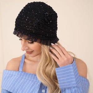 May include: A black crochet hat with a ruffled brim and floral embellishments. The hat is adorned with small, colorful sequins. The person in the image is wearing a blue and white striped off-the-shoulder top.