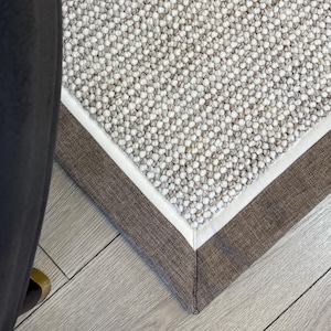 Peut inclure: Un tapis de sol de couleur neutre avec une surface texturée et une bordure marron. Le coin du tapis est visible, mettant en valeur les coutures et le design détaillés. Le tapis est posé sur un sol en bois clair.