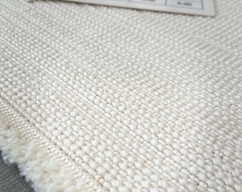 Pode incluir: Close-up de um tapete tecido creme com uma superfície texturizada e borda com franjas. Amostras de cores e um objeto preto são visíveis no fundo, sugerindo um tema de design ou decoração.