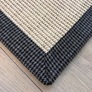 May include: A close-up of a beige and black area rug. The rug has a woven texture with a beige central section and a black and grey patterned border. The rug is rectangular and appears to be made of durable materials.