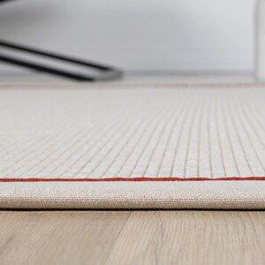 Puede incluir: Primer plano de una alfombra blanca con un fino borde rojo. La alfombra tiene un sutil patr&oacute;n texturizado y est&aacute; doblada en el borde.