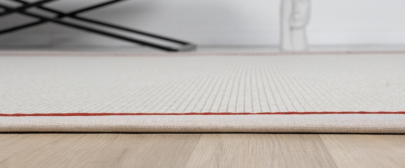 Puede incluir: Primer plano de una alfombra blanca con un fino borde rojo. La alfombra tiene un sutil patr&oacute;n texturizado y est&aacute; doblada en el borde.