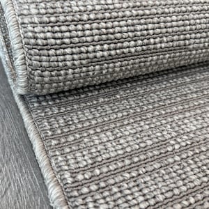 May include: A rolled-up, light gray rug with a textured, woven surface. The rug has a visible border and is partially unrolled, revealing the detailed weave. The neutral color and simple design suggest a focus on functionality and comfort.
