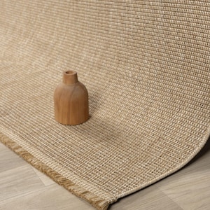 Op de afbeelding: Een beige geweven vloerkleed met een gefranjerde rand en een kleine houten vaas. Het vloerkleed heeft een textuur met een rasterpatroon. De vaas is cilindrisch met een smalle hals. Het vloerkleed ligt op een lichte houten vloer.