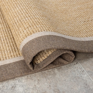 May include: A close-up of a rolled-up natural fibre rug with a brown and white border. The rug is made of a woven material and has a natural, earthy look.