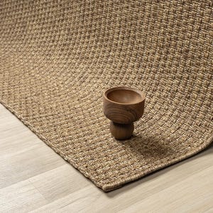 May include: A rectangular rug in shades of tan and brown, with a basketweave pattern. A small, wooden bowl with a rounded interior and a pedestal base sits on the rug. The rug's texture and colour suggest a natural fibre.