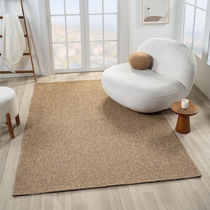 May include: A rectangular, brown area rug with a woven, textured appearance. The rug is placed on a light-colored wooden floor, with a white chair and small wooden side table in the background. A white blanket hangs on a wooden ladder.