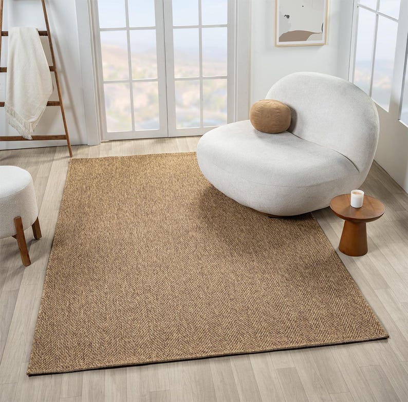 May include: A rectangular, brown area rug with a woven, textured appearance. The rug is placed on a light-colored wooden floor, with a white chair and small wooden side table in the background. A white blanket hangs on a wooden ladder.