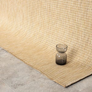 Puede incluir: Una alfombra tejida de color beige y crema con una textura acanalada. Un peque&ntilde;o jarr&oacute;n de cristal gris oscuro se encuentra sobre la alfombra. La alfombra est&aacute; doblada, revelando su reverso y el suelo de baldosas grises.