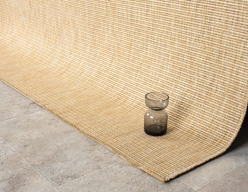 Puede incluir: Una alfombra tejida de color beige y crema con una textura acanalada. Un peque&ntilde;o jarr&oacute;n de cristal gris oscuro se encuentra sobre la alfombra. La alfombra est&aacute; doblada, revelando su reverso y el suelo de baldosas grises.