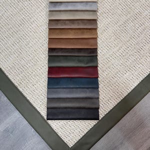 May include: A selection of fabric swatches in various colors, including shades of gray, brown, and blue, arranged on a textured beige surface. The swatches are displayed against a backdrop with a green border, showcasing a range of textures and hues for interior design.