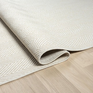 May include: A rolled-up, light beige rug featuring a textured herringbone design. The rug, made from a durable material, sits on a light wood floor. Its neutral colour and pattern make it suitable for various interior styles. The rug's dimensions are in centimeters.