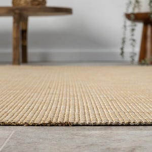 Puede incluir: Primer plano de una alfombra rectangular beige con un dise&ntilde;o texturizado y acanalado. La alfombra est&aacute; hecha de fibras naturales y tiene un aspecto tejido. La alfombra est&aacute; sobre un suelo de baldosas grises.