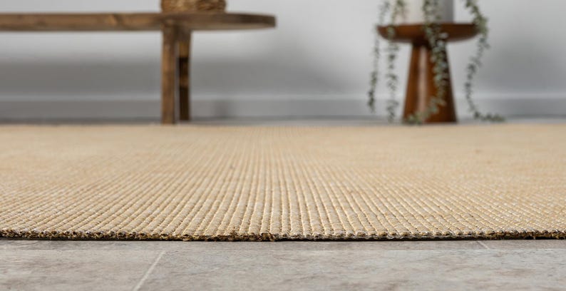 Puede incluir: Primer plano de una alfombra rectangular beige con un dise&ntilde;o texturizado y acanalado. La alfombra est&aacute; hecha de fibras naturales y tiene un aspecto tejido. La alfombra est&aacute; sobre un suelo de baldosas grises.