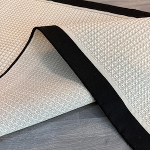 May include: Close-up of a beige and black rug. The rug has a woven pattern with a black border and a lighter beige trim. The rug is folded over, revealing the underside.