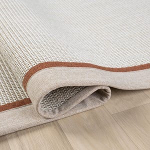 Könnte beinhalten: Nahaufnahme eines beigen Teppichs mit braunem Rand. Der Teppich ist aufgerollt und zeigt die Unterseite des Stoffes.