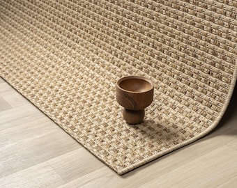 May include: A woven beige rug with a textured pattern. A small, wooden bowl with a pedestal base sits on the rug. The rug has a natural, neutral color palette.