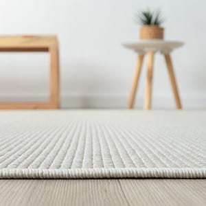May include: Close-up of a light beige rug with a textured, ribbed pattern. The rug's edge is visible, showing a neatly finished border. The background includes a wooden bench and a small side table with a plant.