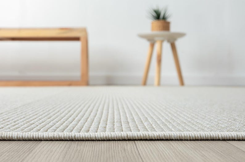 May include: Close-up of a light beige rug with a textured, ribbed pattern. The rug's edge is visible, showing a neatly finished border. The background includes a wooden bench and a small side table with a plant.