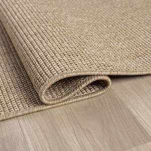 May include: Close-up of a rolled-up, beige woven rug with a textured surface. The rug's edge is visible, showcasing a detailed border. The rug is placed on a light-colored wooden floor, creating a neutral, natural aesthetic.
