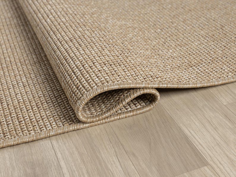 May include: Close-up of a rolled-up, beige woven rug with a textured surface. The rug's edge is visible, showcasing a detailed border. The rug is placed on a light-colored wooden floor, creating a neutral, natural aesthetic.