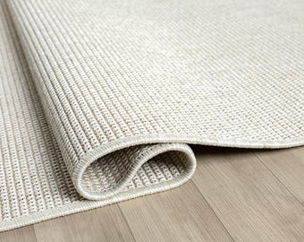May include: A rolled-up, neutral-toned area rug with a textured weave. The rug's edge is neatly finished with a subtle border. The rug is placed on a light-colored wooden floor, showcasing its texture and design.