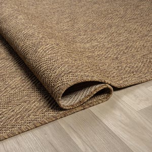 May include: A rolled-up, textured brown rug with a woven pattern. The rug's edge is visible, revealing a lighter-colored underside. The rug is placed on a light-colored wooden floor, creating a contrast in textures and tones.