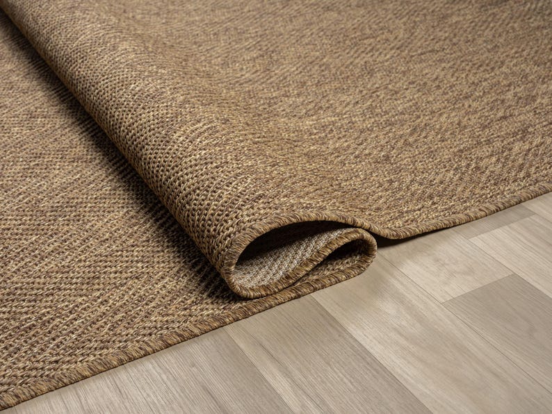 May include: A rolled-up, textured brown rug with a woven pattern. The rug's edge is visible, revealing a lighter-colored underside. The rug is placed on a light-colored wooden floor, creating a contrast in textures and tones.