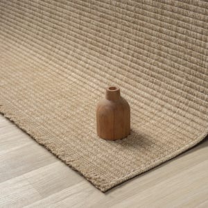 May include: A beige woven rug with a ribbed texture and a fringed edge. A small, wooden vase with a rounded body and a narrow neck sits on the rug. The rug is made of a natural fibre.