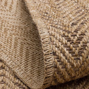 Könnte beinhalten: Nahaufnahme eines gewebten Teppichs in Naturtönen. Der Teppich hat ein strukturiertes Fischgrätmuster in Beige- und Brauntönen. Der Rand des Teppichs ist sichtbar und zeigt einen detaillierten, genähten Rand.
