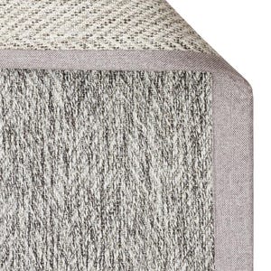 Puede incluir: Primer plano de una sección de una alfombra tejida con un patrón de espiga en tonos grises y blancos. La alfombra tiene un borde gris claro. La esquina de la alfombra está ligeramente enrollada, revelando la parte inferior.