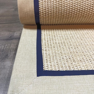 Könnte beinhalten: Eine aufgerollte, beige gewebte Matte mit einem marineblauen Rand. Die Matte hat ein strukturiertes Aussehen aus Naturfasern mit einem durchgehenden marineblauen Rand. Die Matte ist teilweise aufgerollt und gibt die Unterseite frei.