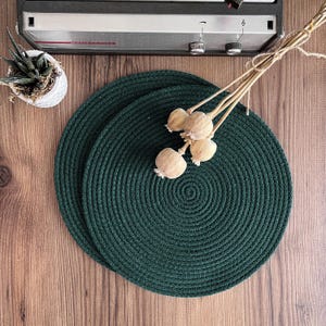 Può includere: Due sottopiatti circolari verde scuro in materiale tessuto sono esposti su una superficie di legno. Sono presenti anche baccelli di semi secchi e una piccola pianta succulenta in vaso. Sullo sfondo è visibile uno stereo vintage.