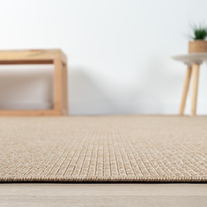 May include: Close-up of a beige woven rug with a textured, rectangular pattern. The rug is on a light-colored wooden floor. In the background, a wooden bench and a small side table with a plant are visible.