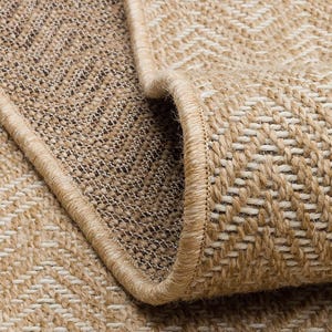 Op de afbeelding: Close-up van een opgerold beige en bruin tapijt met een geweven visgraatpatroon. Het tapijt heeft een beige rand en een getextureerd oppervlak, met een natuurlijk, neutraal kleurenpalet.
