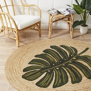May include: A round jute rug with a large green leaf design. The rug is natural in color and has a woven texture.