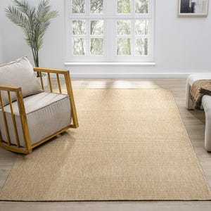 Puede incluir: Una alfombra rectangular beige con una textura tejida, colocada sobre un suelo de madera de color claro. A la izquierda hay una mecedora de madera con un coj&iacute;n y una almohada beiges. Una ventana blanca y una planta est&aacute;n en el fondo.