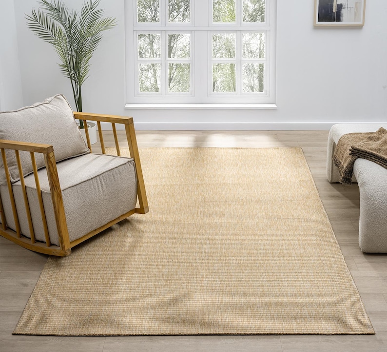 Puede incluir: Una alfombra rectangular beige con una textura tejida, colocada sobre un suelo de madera de color claro. A la izquierda hay una mecedora de madera con un coj&iacute;n y una almohada beiges. Una ventana blanca y una planta est&aacute;n en el fondo.