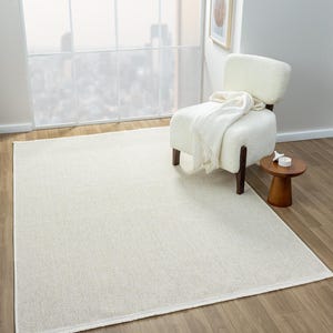 May include: A cream-colored rectangular area rug with a woven texture. The rug is placed on a hardwood floor, with a white chair and small wooden side table in the background. A white blanket is draped over the chair.