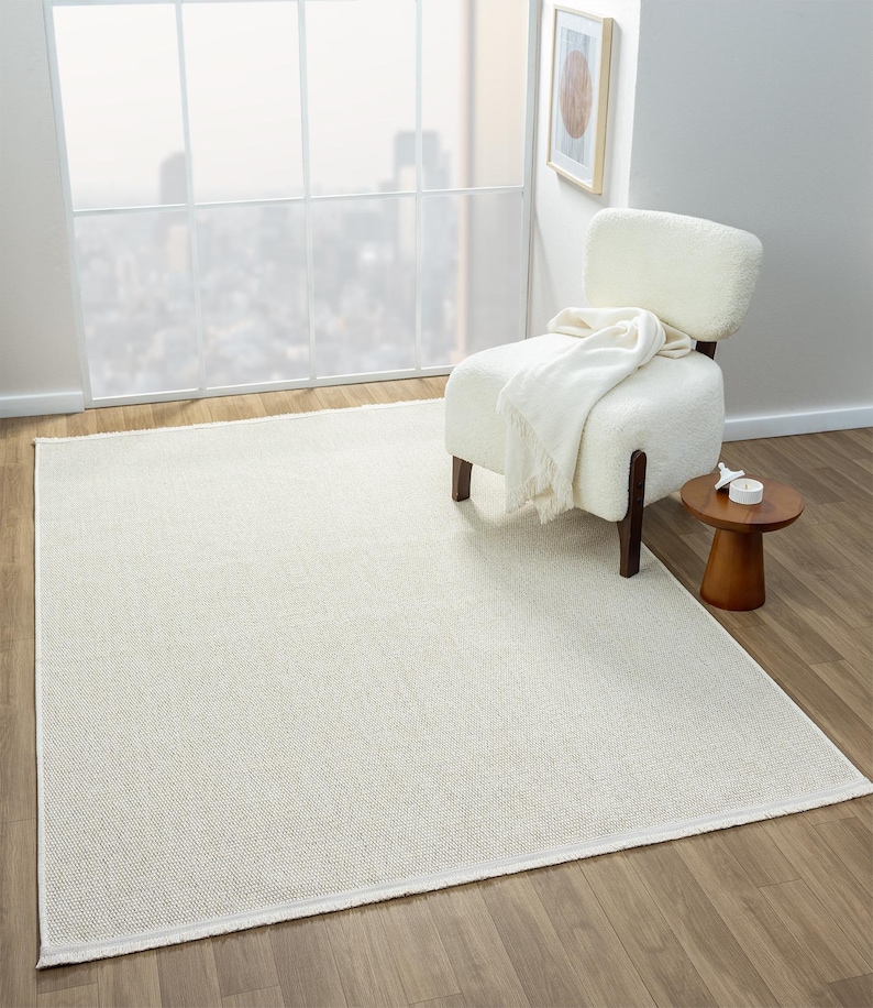 May include: A cream-colored rectangular area rug with a woven texture. The rug is placed on a hardwood floor, with a white chair and small wooden side table in the background. A white blanket is draped over the chair.