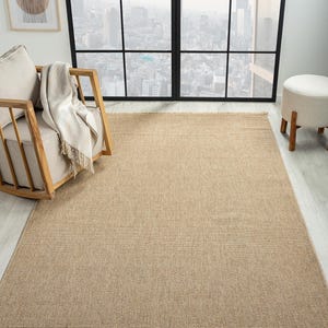 May include: A rectangular, beige woven rug with a textured surface. The rug has a fringe detail along the shorter edges. The rug is placed on a light-colored wooden floor. A wooden chair with a cream-colored cushion and a blanket is in the background.