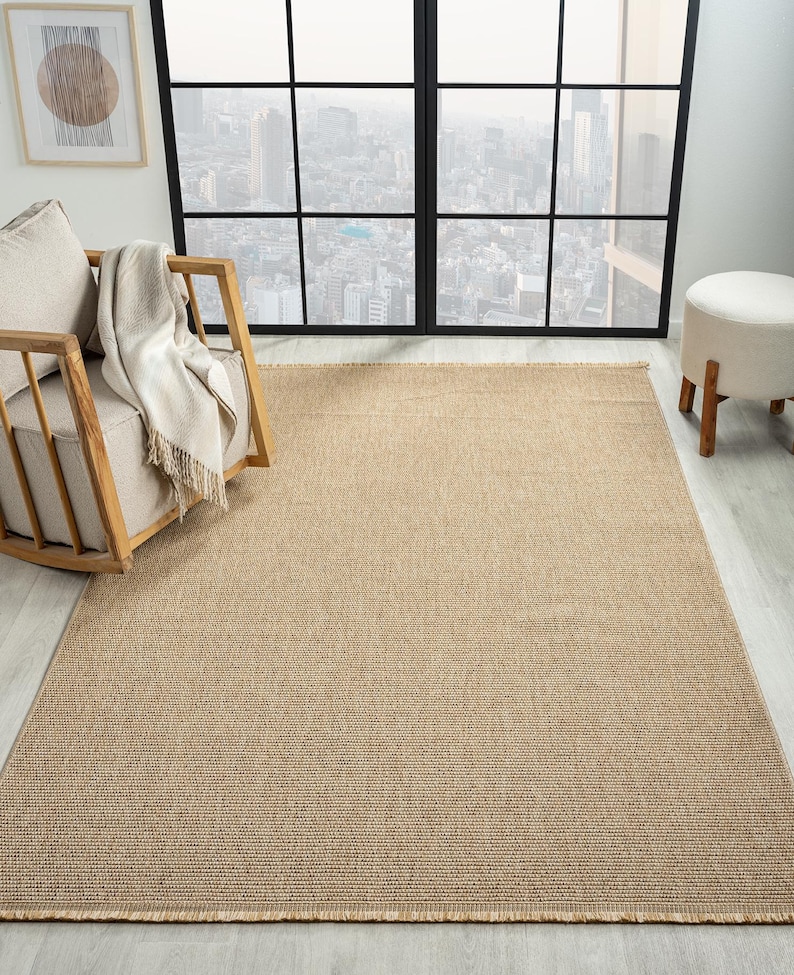 May include: A rectangular, beige woven rug with a textured surface. The rug has a fringe detail along the shorter edges. The rug is placed on a light-colored wooden floor. A wooden chair with a cream-colored cushion and a blanket is in the background.