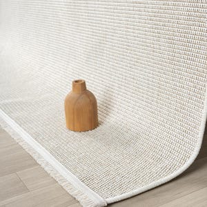 May include: A neutral-toned area rug with a textured weave and a fringed edge. A small, wooden decorative vase sits on the rug. The rug's color is a light beige, and the fringe is white.