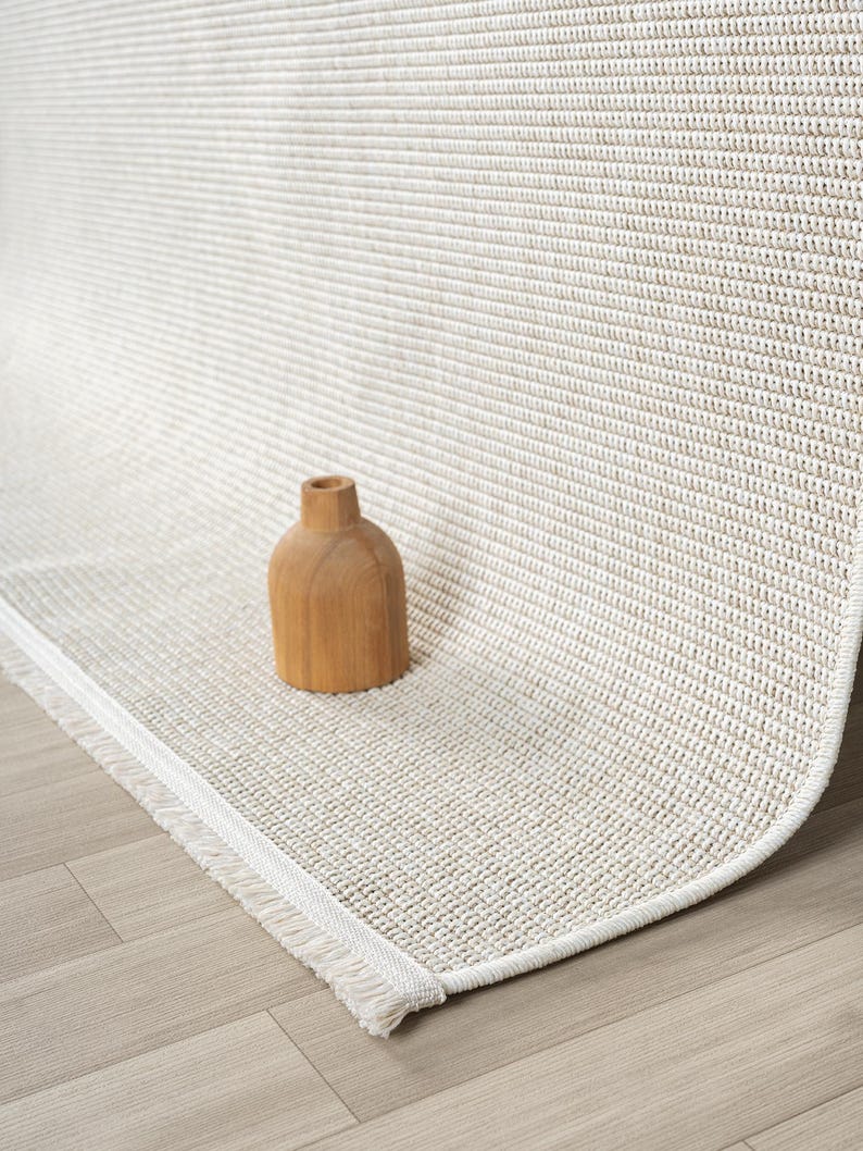 May include: A neutral-toned area rug with a textured weave and a fringed edge. A small, wooden decorative vase sits on the rug. The rug's color is a light beige, and the fringe is white.