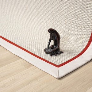 Puede incluir: Una alfombra blanca con un borde rojo y una textura tejida. La alfombra tiene un respaldo de tela blanca y est&aacute; doblada por la mitad. Una peque&ntilde;a figurita marr&oacute;n oscura de una persona est&aacute; sentada en la alfombra.