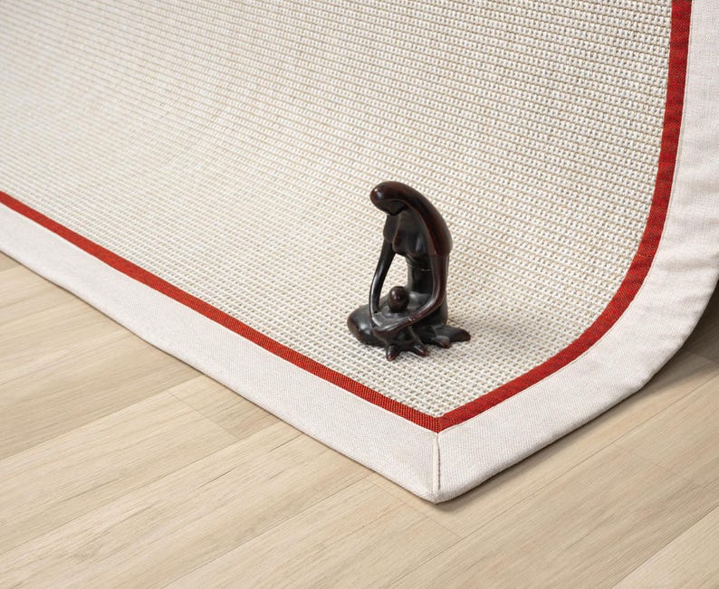 Puede incluir: Una alfombra blanca con un borde rojo y una textura tejida. La alfombra tiene un respaldo de tela blanca y est&aacute; doblada por la mitad. Una peque&ntilde;a figurita marr&oacute;n oscura de una persona est&aacute; sentada en la alfombra.
