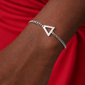 May include: A silver-colored bracelet featuring a triangular charm. The bracelet has a chain link design and is worn on a wrist against a red background. The triangle is a simple, geometric shape.