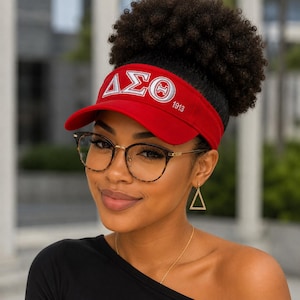 May include: A red visor with white Greek letters and the number 1913. The visor is worn by a person wearing glasses and gold earrings. The visor has a curved brim and a band that fits around the head.